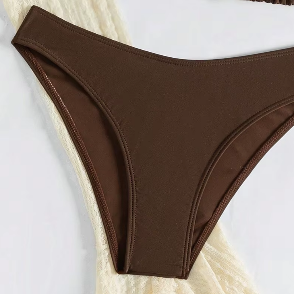 girlzhood Saint Tropez Muse Set Bikini-Slip in Braun auf einem cremefarbenen, strukturierten Stoffhintergrund.